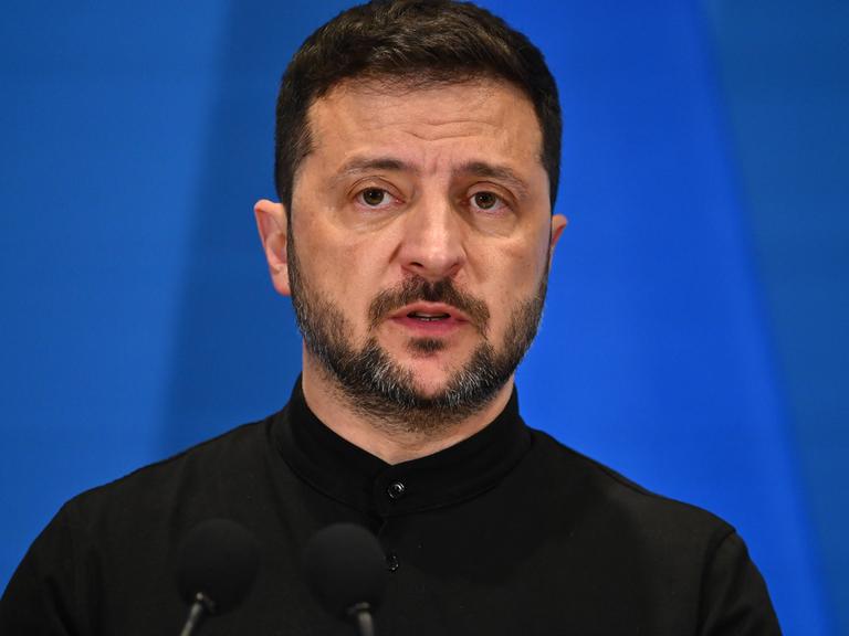 Wolodymyr Selenskyj steht auf einem Podium vor blauem Hintergrund.