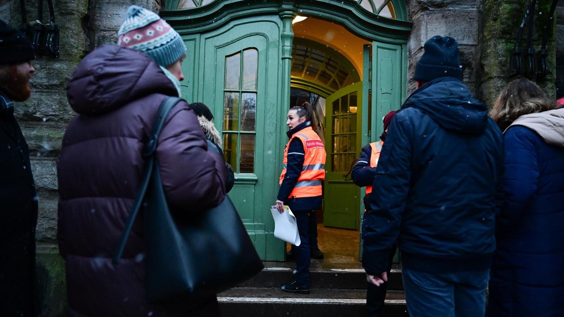 Eine Frau mit einer Uniform macht die Tür zu einem Gebäude auf. Davor stehen wartende Menschen. Es ist dunkel. Eine Frau mit einer Uniform macht die Tür zu einem Gebäude auf. Davor stehen wartende Menschen. Es ist dunkel.
