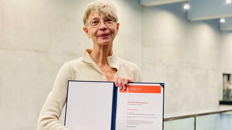 Eine Frau mit kurzen Haaren und Brille hält ein Zertifikat mit einem Medienpreis in die Kamera Eine Frau mit kurzen Haaren und Brille hält ein Zertifikat mit einem Medienpreis in die Kamera