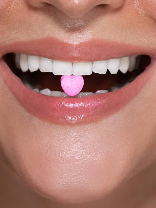 Symbolbild: Eine Frau hat eine herzförmige Tablette zwischen ihren Zähnen, die die illegale Partydroge MDMA symbolisieren soll.