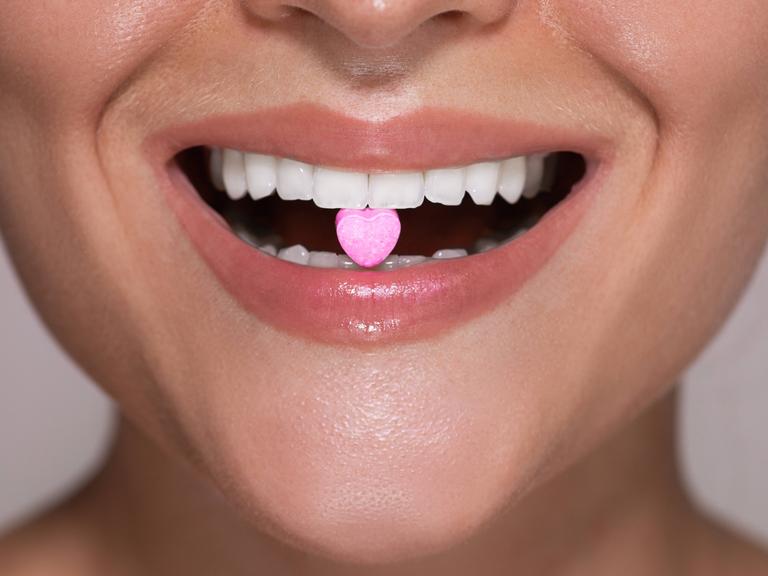 Symbolbild: Eine Frau hat eine herzförmige Tablette zwischen ihren Zähnen, die die illegale Partydroge MDMA symbolisieren soll.