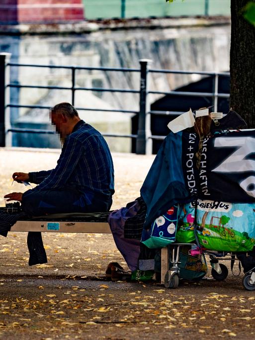 Ein obdachloser Mensch sitzt auf einer Bank im Schatten der Baeumen im Lustgarten auf der Museumsinsel in Berlin.