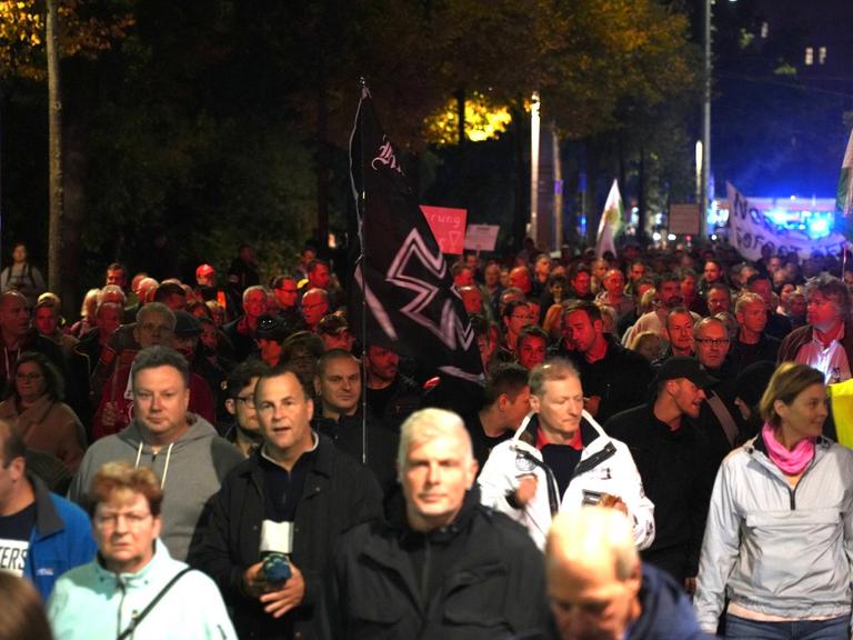 Viele Menschen demonstrieren auf einer Straße, in der Mitte ist eine alte Reichskriegsflagge mit schwarzem Kreuz zu sehen.