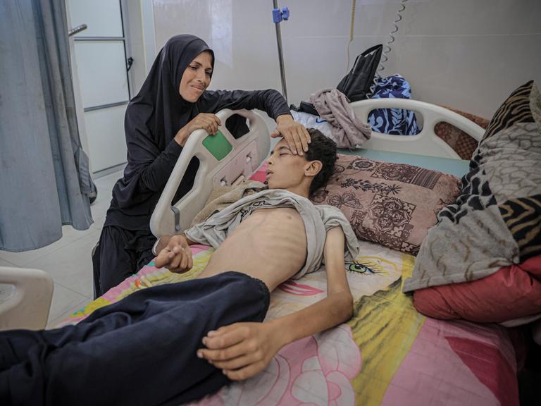 Ein stark abgemagerter Jugendlicher liegt im Al-Schifa-Krankenhaus in Gaza-Stadt in einem Krankenbett und wartet auf Behandlung. Seine Mutter kümmert sich um ihn.