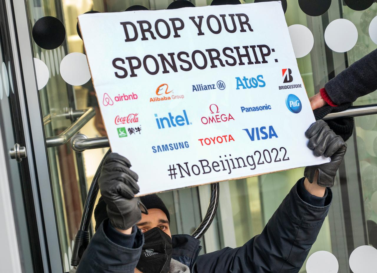 Vor dem Eingang einer Niederlassung der Allianz Versicherung in Berlin steht ein Aktivist mit einem Plakat mit der Aufschrift "Drop your Sponsorship", darunter die Logos von airbnb, Alibaba, Allianz, Atos, Bridgestone, CocaCola, intel, omega, panasonic, P&G, Samsung, Toyota und Visa.