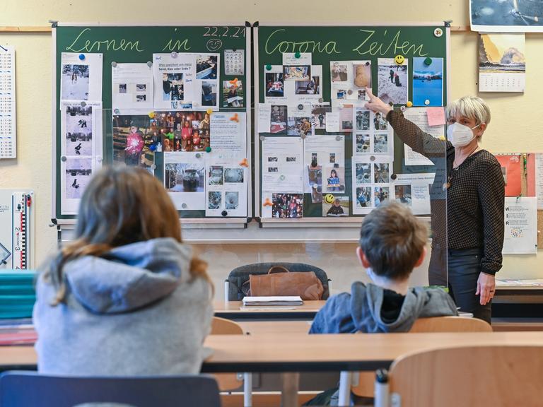 Die Lehrerin einer 4. Klasse an der Grundschule "Am Mühlenfließ" in Frankfurt (Oder), unterrichtet zwei SchülerInnen in einem Klassenzimmer. Auf der Tafal steht "Lernen in Corona Zeiten".