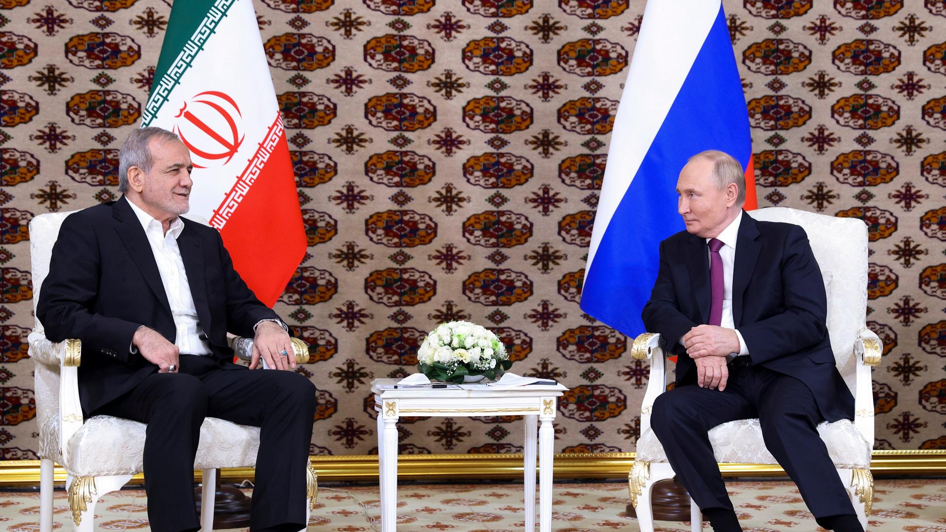 Russlands Präsident Putin und der iranische Präsident Peseschkian unterhalten sich. Sie sitzen auf weißen Stühlen, im Hintergrund die jeweilige Landesflaggen.