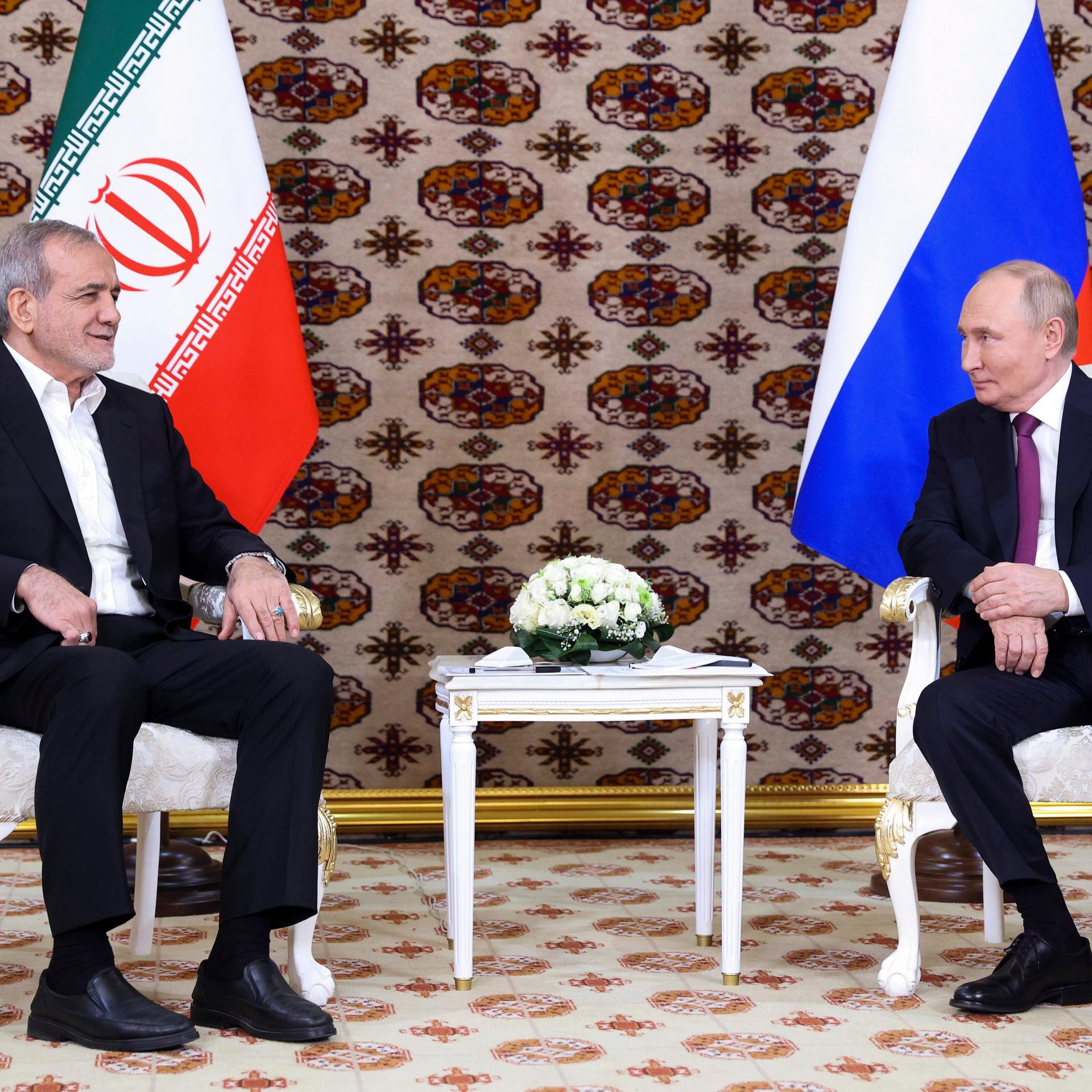 Russlands Präsident Putin und der iranische Präsident Peseschkian unterhalten sich. Sie sitzen auf weißen Stühlen, im Hintergrund die jeweilige Landesflaggen.