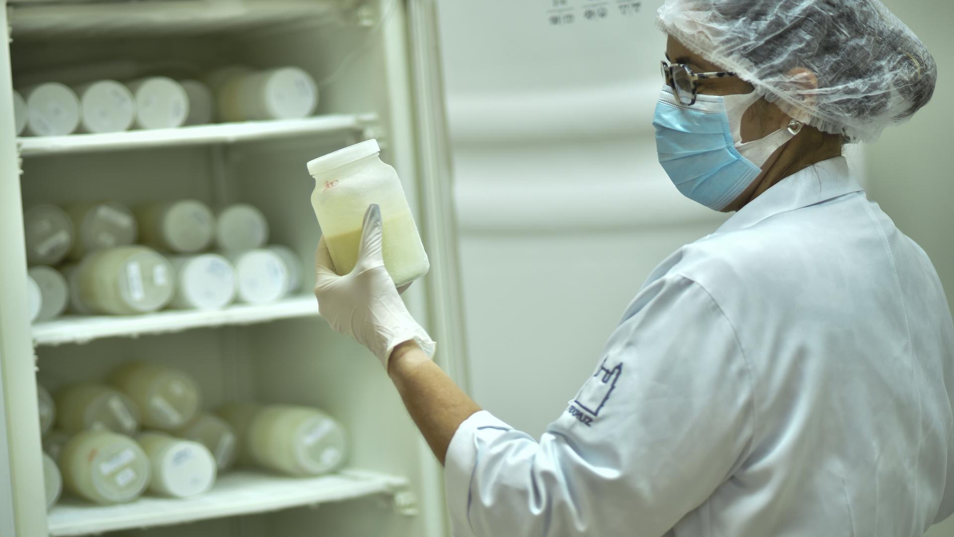 Eine Krankenschwester ordnet Flaschen mit Muttermilch in einem Kühlschrank. Sie trägt einen Haarschutz und einen Mund-Nasen-Schutz. Eine Krankenschwester ordnet Flaschen mit Muttermilch in einem Kühlschrank. Sie trägt einen Haarschutz und einen Mund-Nasen-Schutz.