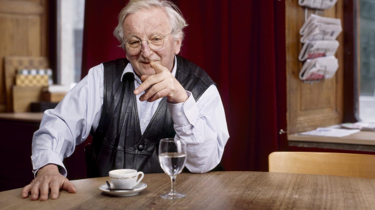 Peter Bichsel sitzt lächelnd am Tisch und zeigt in die Kamera.