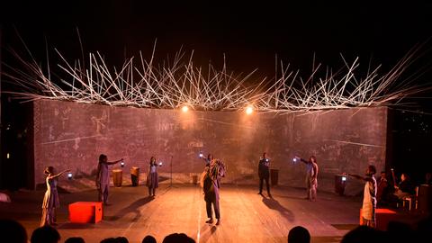 Eine Schauspielertruppe steht auf einer spärlich beleuchteten Gruppe, die kleine Lampen in der Hand halten. Dabei kreist die Gruppe einen Mann mit einem Sack auf der Schulter ein.