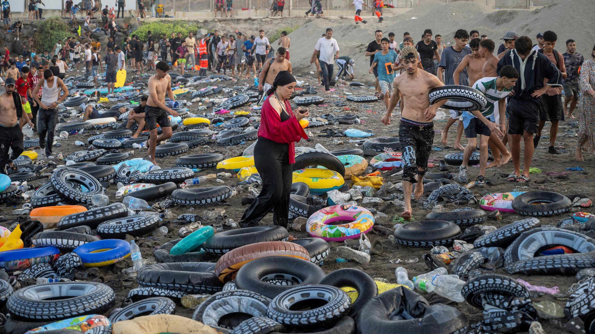 Am Strand von Ceuta liegen Hunderte bunte Plastik-Schwimmringe auf dem Sand. Darüber steigen dutzende Menschen in nasser Kleidung. Im Hintergrund steht ein meterhoher Grenzzaun.