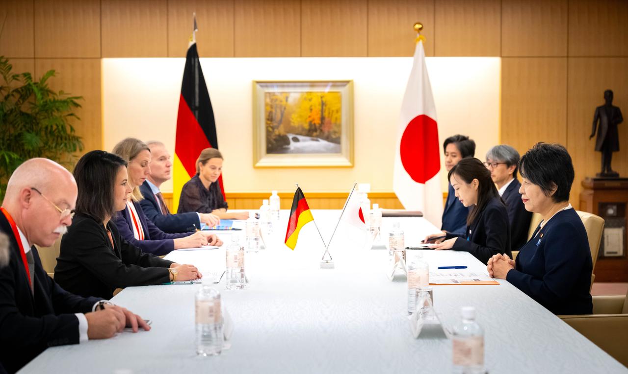 Mehrere Menschen sitzen an einem langen, weißen Tisch. Im Hintergrund sind die deutsche und die japanische Flagge zu sehen.