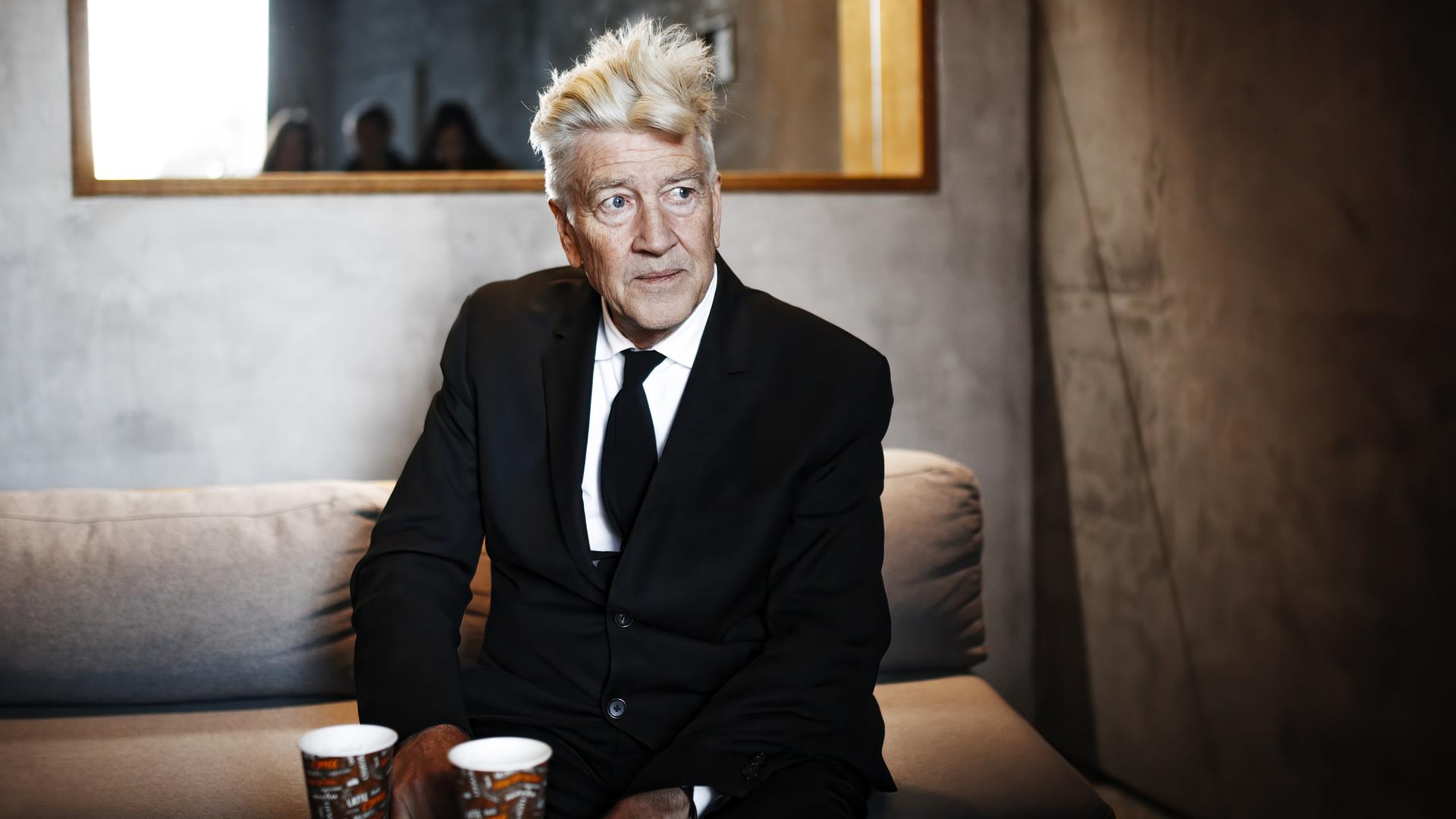 David Lynch sitzt 2017 bei einem Pressetermin auf einem Sofa und schaut nach rechts.