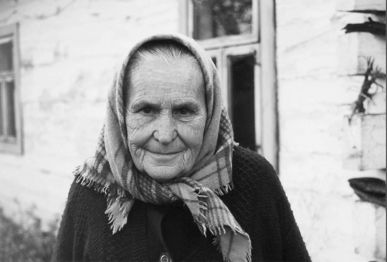Das Foto zeigt eine sehr alte Frau mit faltigem Gesicht. Sie steht in einem dörflichen Garten und trägt ein Kopftuch. Sie blickt direkt in die Kamera.