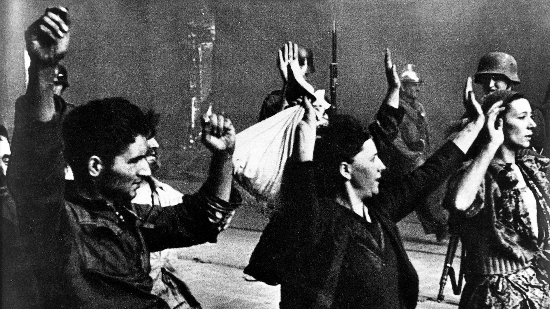 Historische Aufnahme von einem Mann und zwei Frauen mit erhobenen Armen, die von der SS-Polizei abgeführt werden.