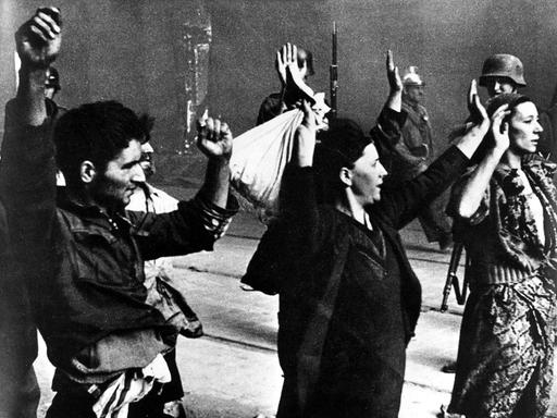 Historische Aufnahme von einem Mann und zwei Frauen mit erhobenen Armen, die von der SS-Polizei abgeführt werden.