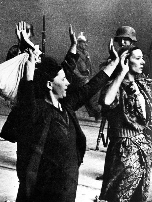 Historische Aufnahme von einem Mann und zwei Frauen mit erhobenen Armen, die von der SS-Polizei abgeführt werden.