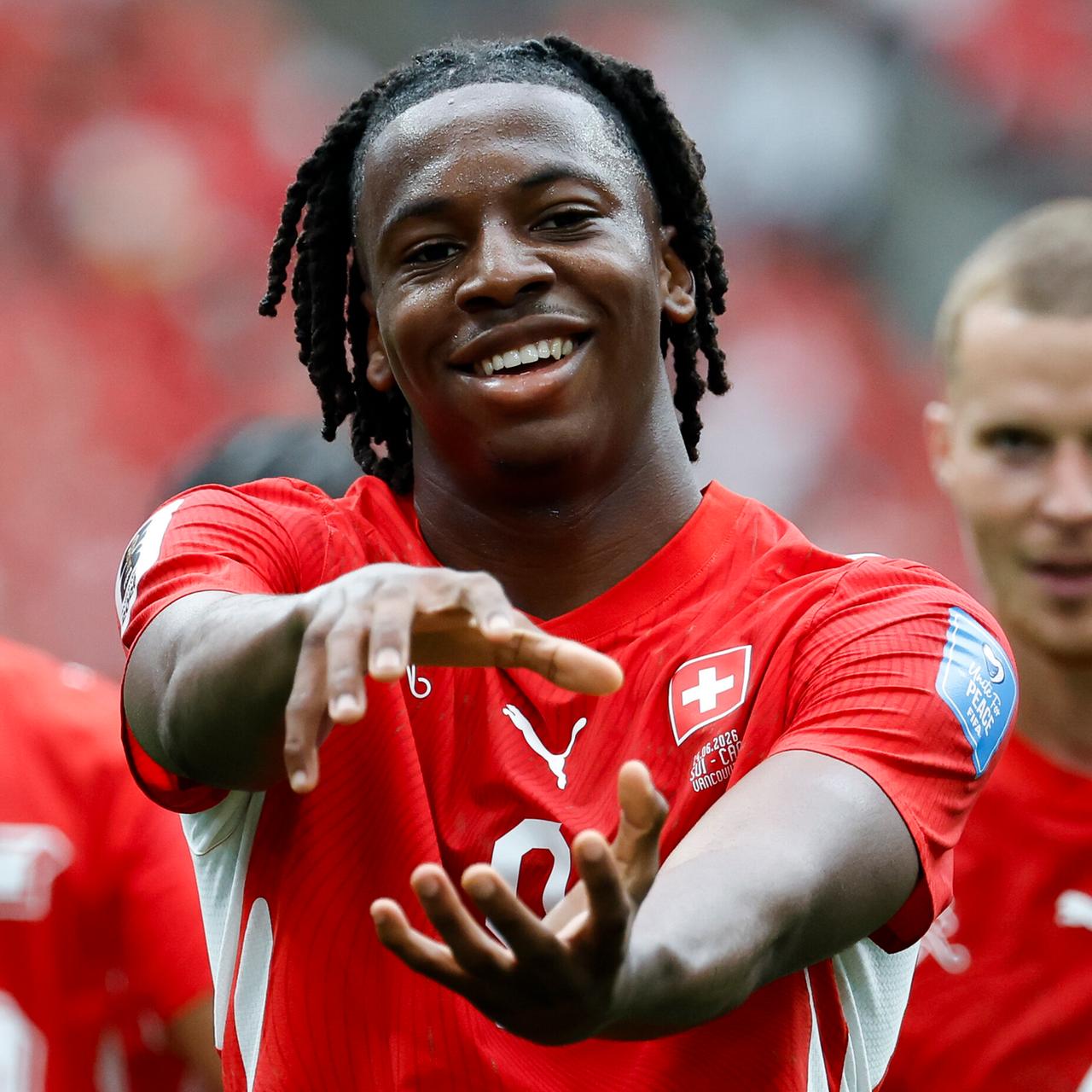 Manzambi im roten Trikot hält lächelnd seine Hände so vor sich, als hielte er eine Kugel. Dahinter unscharf zwei Mitspieler.