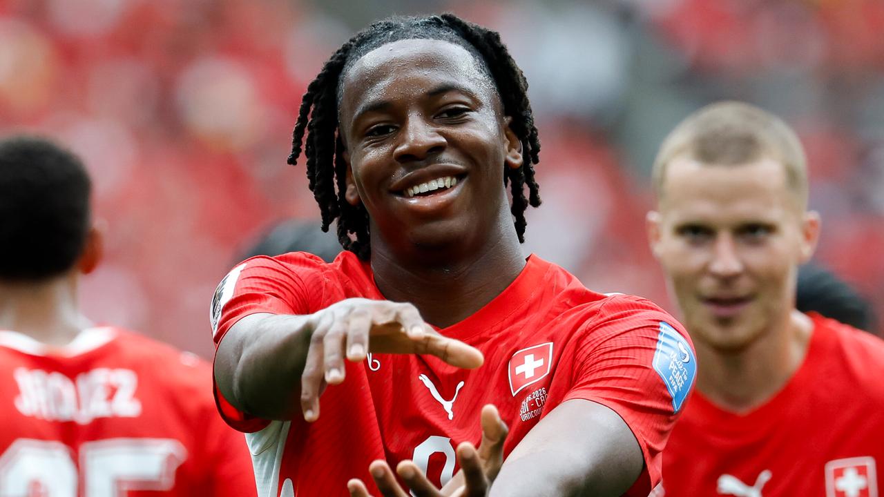 Manzambi im roten Trikot hält lächelnd seine Hände so vor sich, als hielte er eine Kugel. Dahinter unscharf zwei Mitspieler.