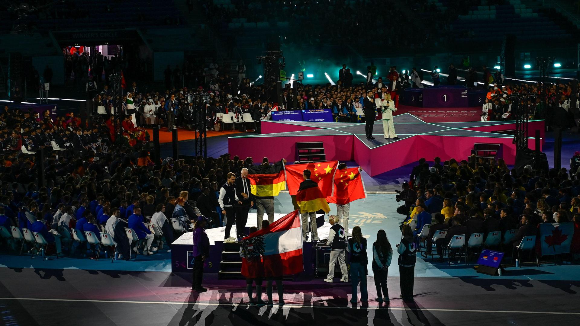 In einem Stadion stehen Menschen mit der deutschen Flagge und andere mit der chinesischen Flagge auf einem Podium. Davor zwei Menschen mit der österreichischen Flagge.