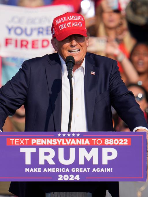 Donald Trump steht hinter einem Rednerpult mit seinem Namen und trägt eine rota "Make America Great Again"-Kappe.