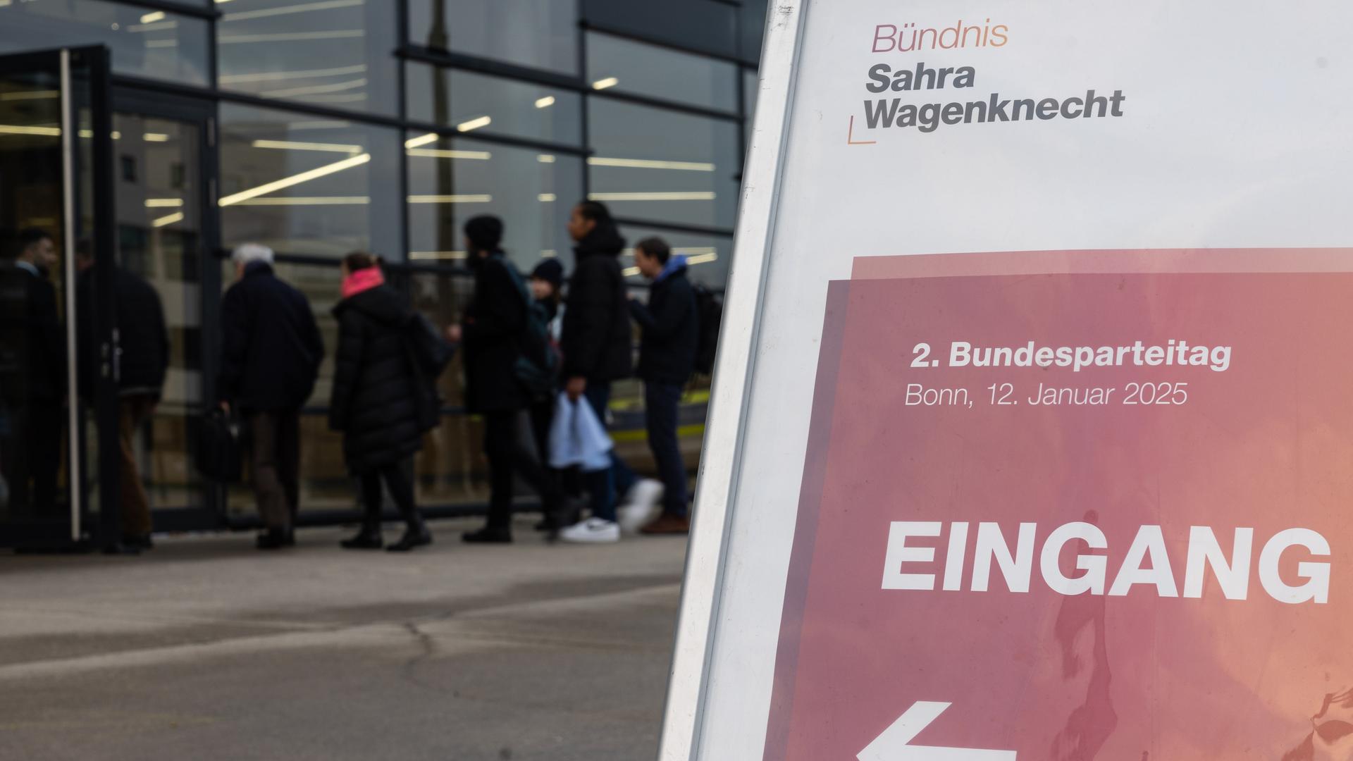 Hinter einem Aufsteller mit der Aufschrift "Bündnis Sahra Wagenknecht" sind Menschen zu sehen, die eine Veranstaltungshalle durch eine gläserne Drehtür betreten wollen. Hinter einem Aufsteller mit der Aufschrift "Bündnis Sahra Wagenknecht" sind Menschen zu sehen, die eine Veranstaltungshalle durch eine gläserne Drehtür betreten wollen.