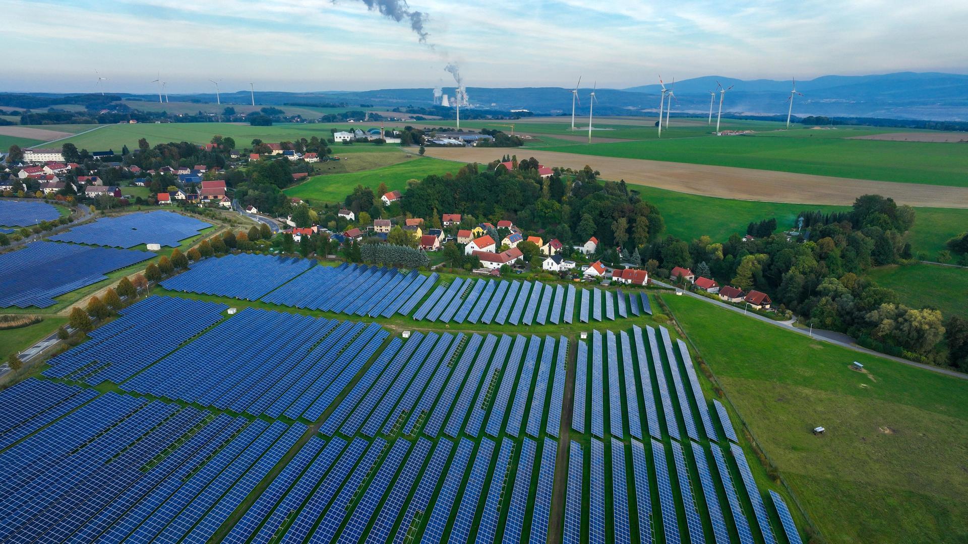 Ein Solarfeld liegt auf einer grünen Wiese, direkt im Anschluss dahinter ein Dorf. Im Hintergrund Wiesen mit Windkrafträdern und Wälder