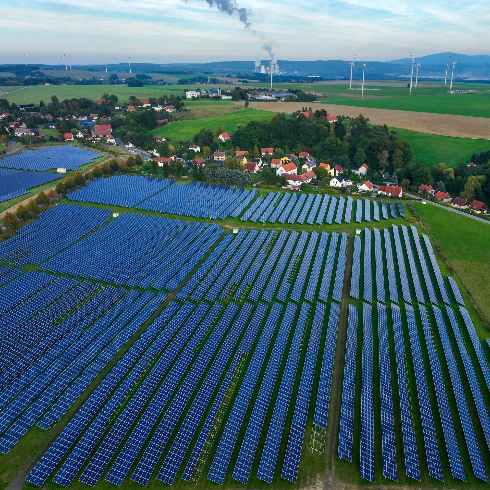 Ein Solarfeld liegt auf einer grünen Wiese, direkt im Anschluss dahinter ein Dorf. Im Hintergrund Wiesen mit Windkrafträdern und Wälder