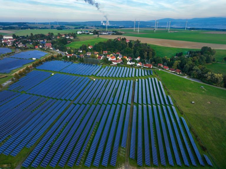 Ein Solarfeld liegt auf einer grünen Wiese, direkt im Anschluss dahinter ein Dorf. Im Hintergrund Wiesen mit Windkrafträdern und Wälder