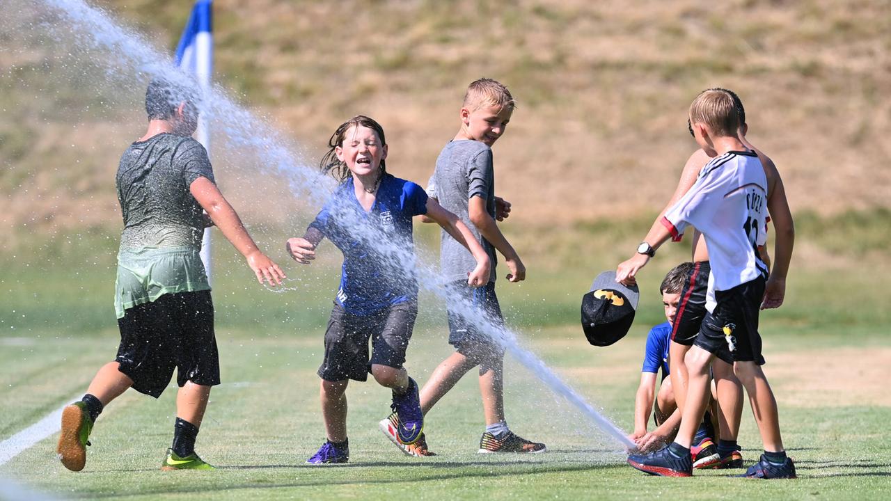 Eine Gruppe von fußballspielenden Jungen erfrischt sich an einem heißen Sommertag auf dem Spielfeld mit Wasser aus einem Rasensprenger.
