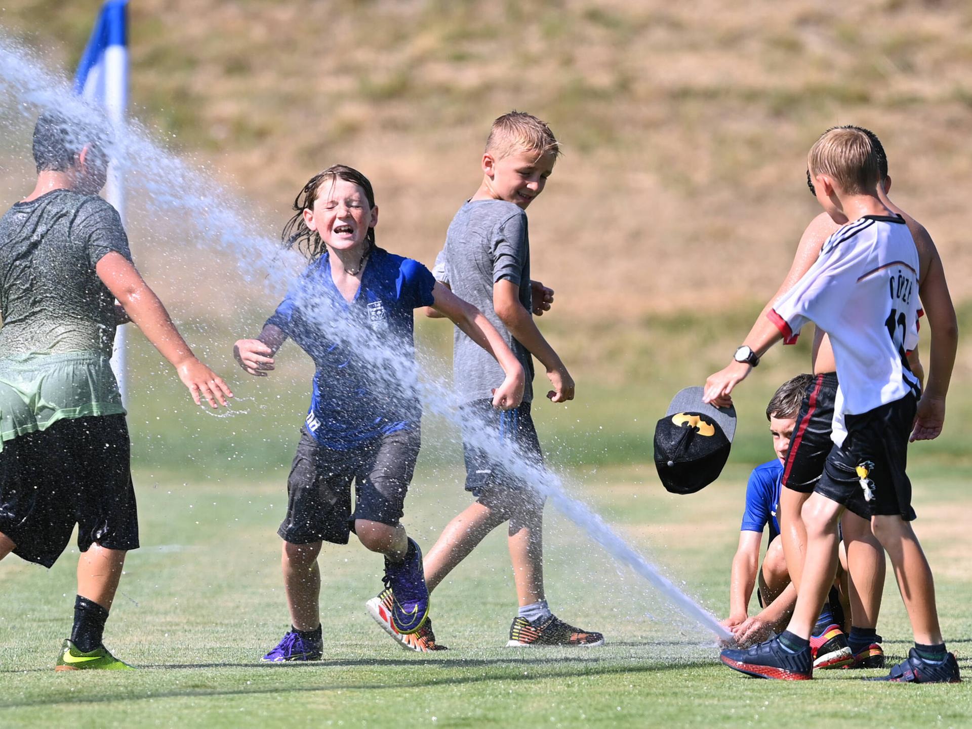 Eine Gruppe von fußballspielenden Jungen erfrischt sich an einem heißen Sommertag auf dem Spielfeld mit Wasser aus einem Rasensprenger.
