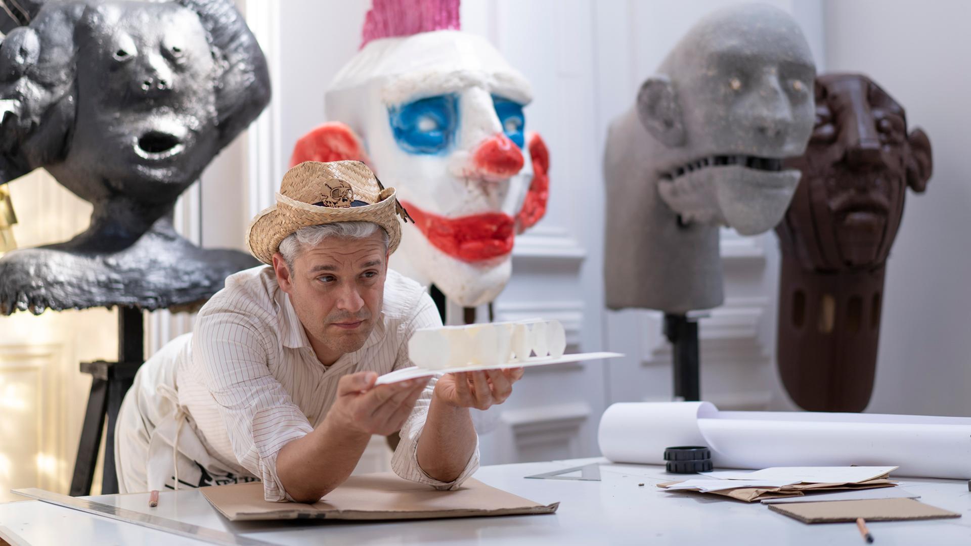 Tobias Binderberger stützt sich in seinem Atelier auf einen Tisch und hält ein Kunstobjekt in der Hand. Auf dem Kopf trägt er einen Strohhut.