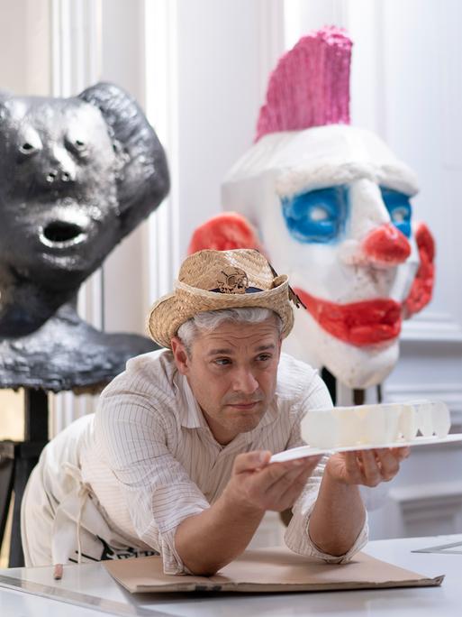 Tobias Binderberger stützt sich in seinem Atelier auf einen Tisch und hält ein Kunstobjekt in der Hand. Auf dem Kopf trägt er einen Strohhut.