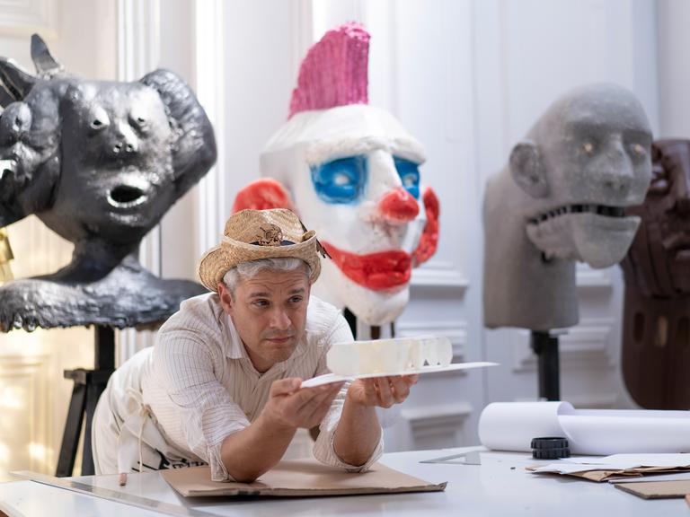 Tobias Binderberger stützt sich in seinem Atelier auf einen Tisch und hält ein Kunstobjekt in der Hand. Auf dem Kopf trägt er einen Strohhut.