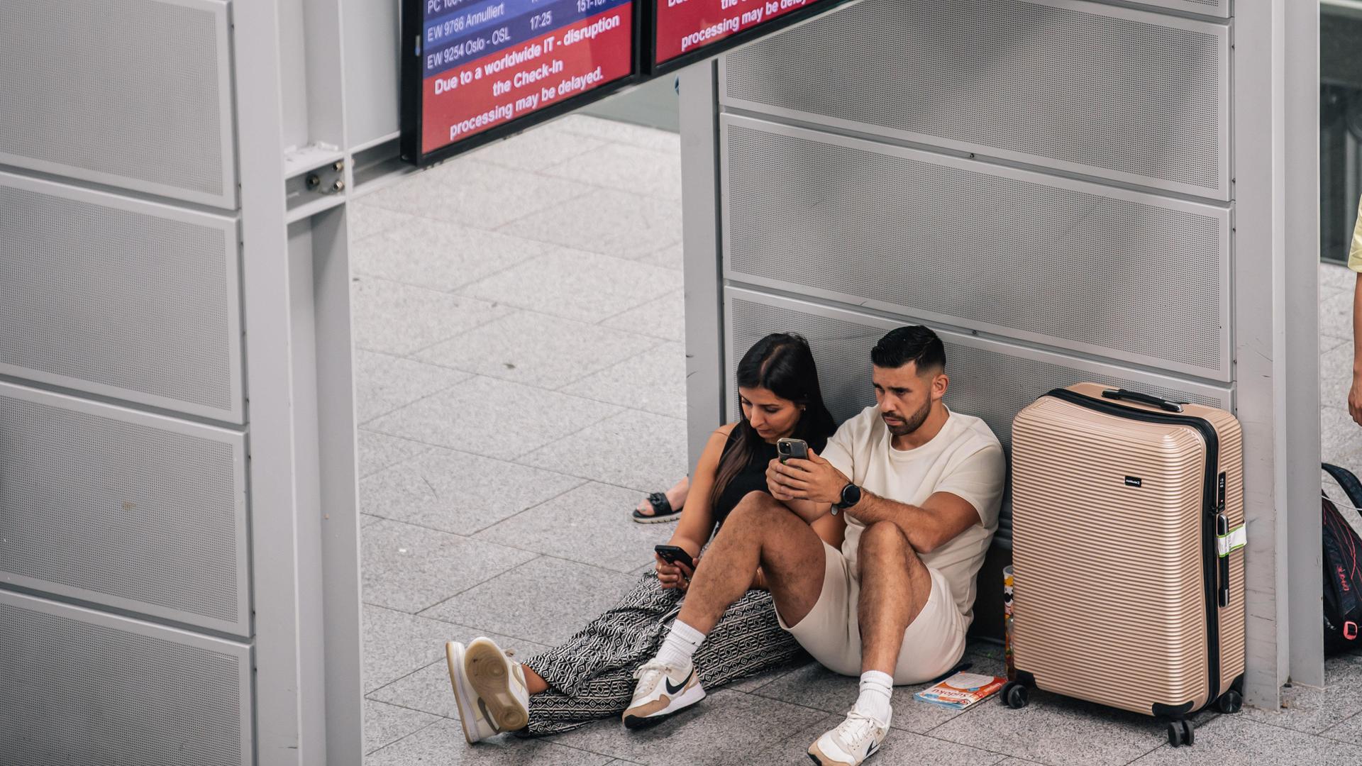 Eine Frau und ein Mann lehnen neben ihrem Koffer an einer Säule in einem Flughafen und gucken auf ihre Handys.