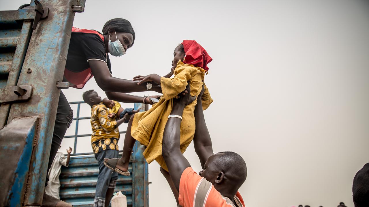 Ein Kind wird von einem Mann in einen Lastwagen gehoben. Eine Frau nimmt es entgegen. Die Menschen fliehen vor dem Krieg im Sudan - von Joda aus erst in ein Durchgangslager in Renk im Südsudan.