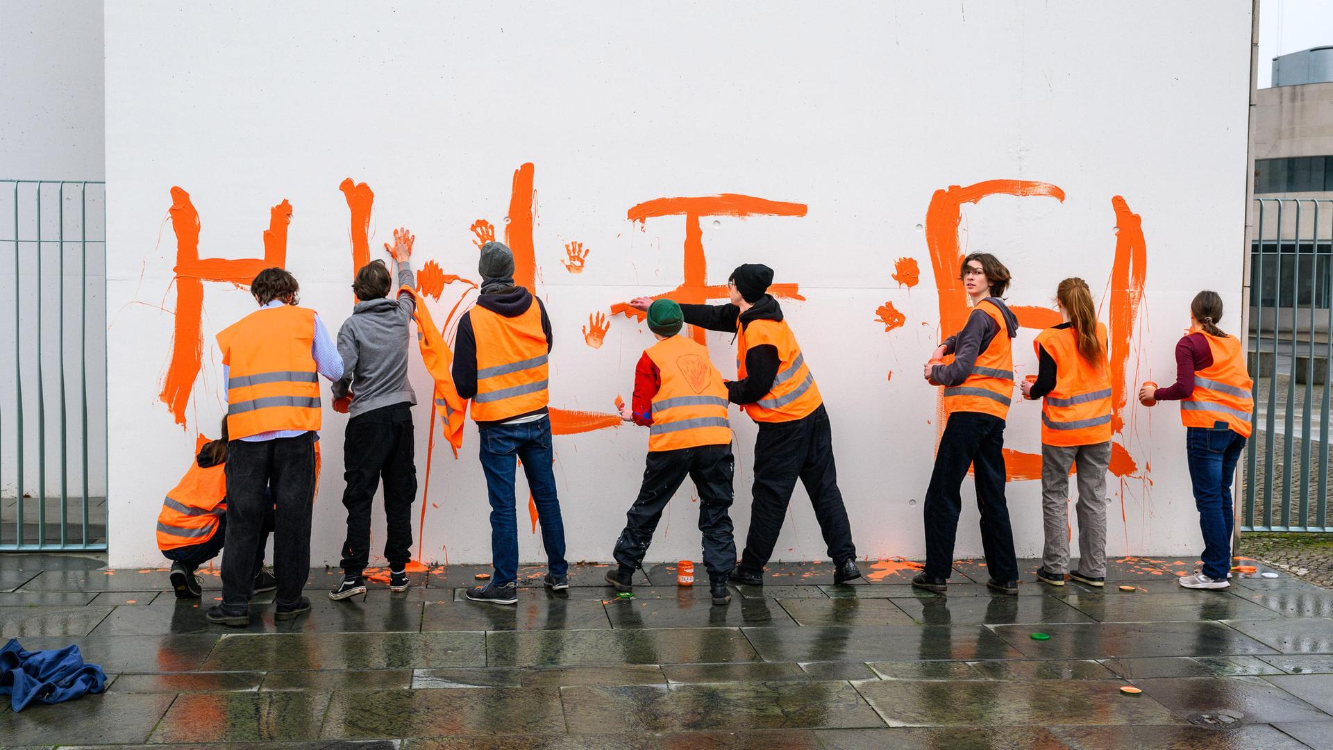 Jugendliche Aktivisten und Aktivistinnen der Letzten Generation schreiben mit orangener Farbe "Hilfe! Eure Kinder" an eine Mauer rund um das  Bundeskanzleramt in Berlin. Im März 2024