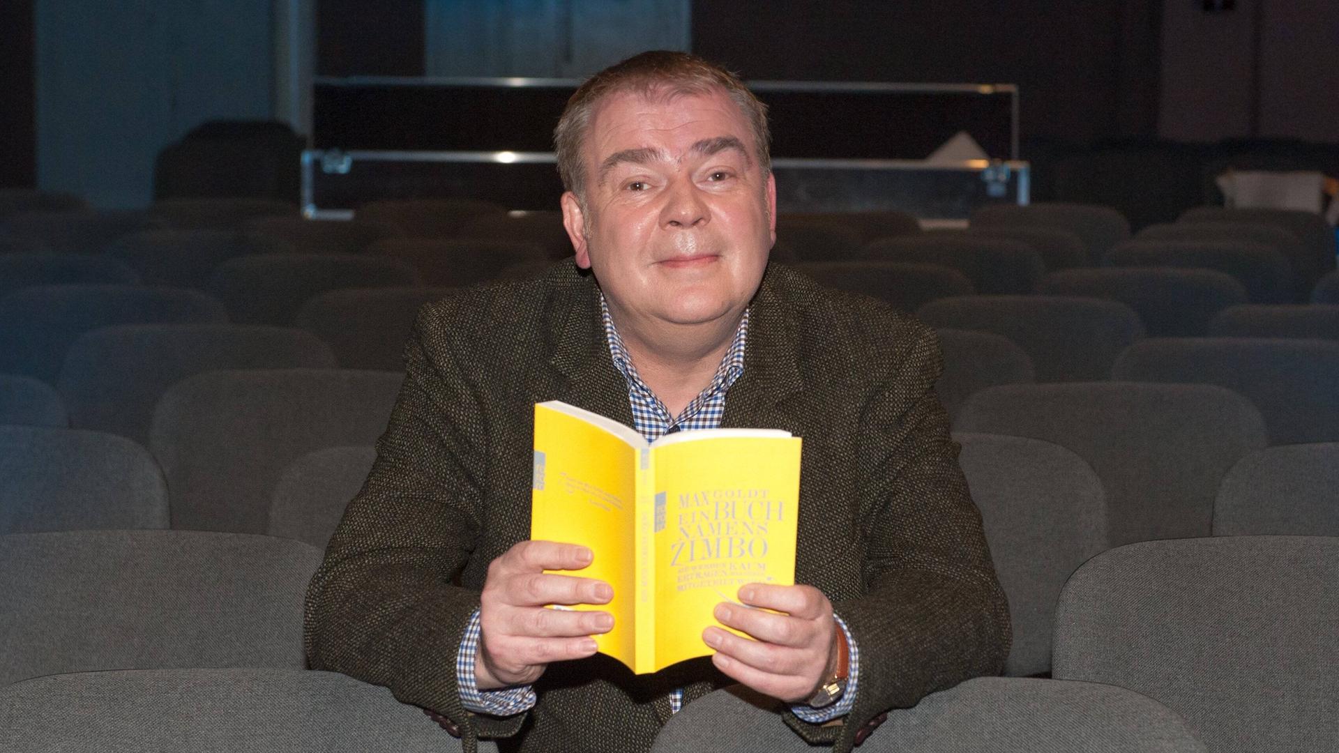 Max Goldt sitzt mit einem gelben Buch in der Hand