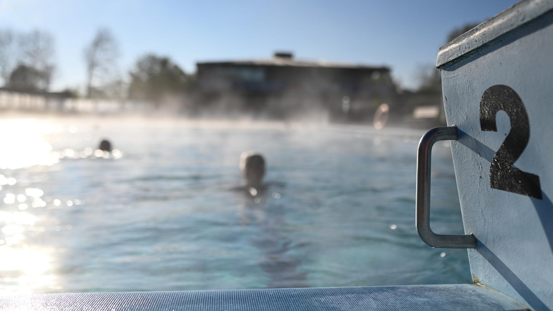 Zwei Menschen schwimmen im Becken eines Freibades, auf dem Nebelschwaden liegen. Im Vordergrund ist ein Startblock zu sehen.