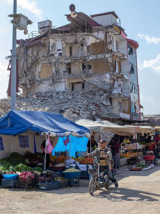 Hinter einem Straßenmarkt erhebt sich die Ruine eines im Erdbeben zerstörten Wohnhauses.