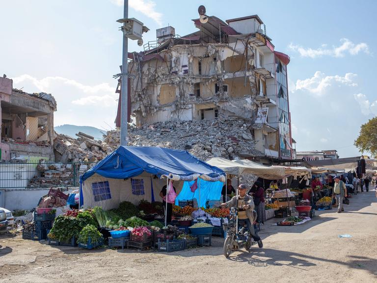 Hinter einem Straßenmarkt erhebt sich die Ruine eines im Erdbeben zerstörten Wohnhauses.