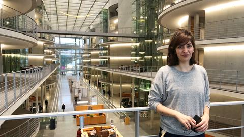 Ein junge Frau mit halblangen dunklen Haaren, in Jeans und grauem Pullover, steht in der Halle des Deutschen Bundestages.