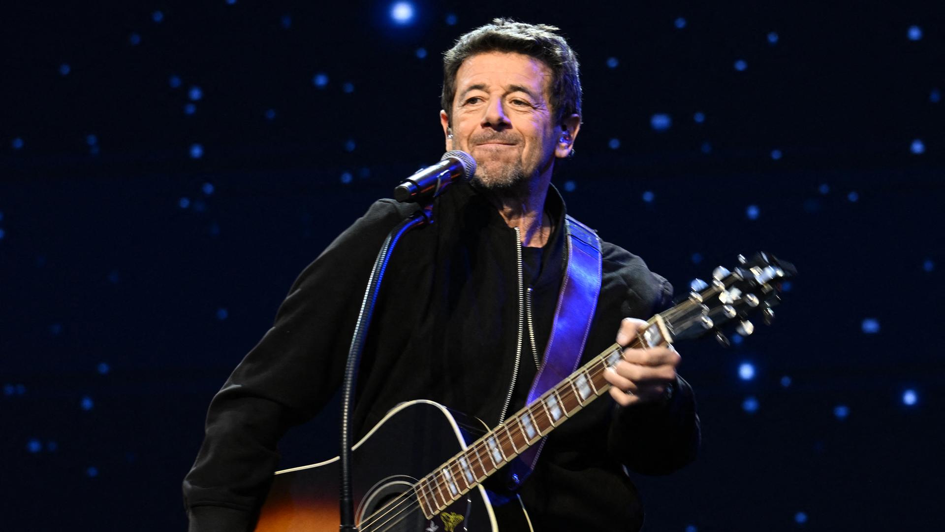 Patrick Bruel steht mit einer Gitarre auf einer Bühne vor dunklem Hintergrund mit sternenartigen Lichtern. 