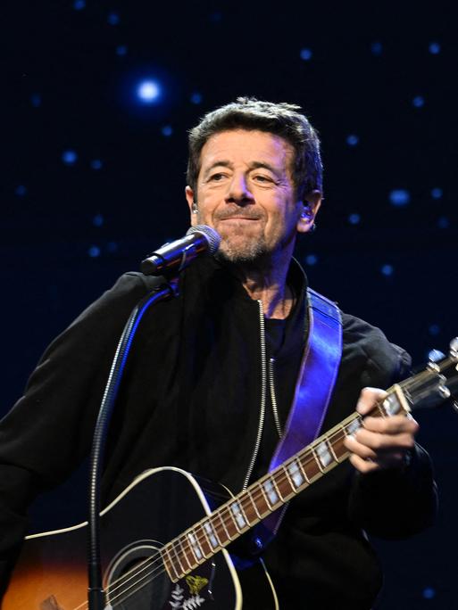 Patrick Bruel steht mit einer Gitarre auf einer Bühne vor dunklem Hintergrund mit sternenartigen Lichtern. 