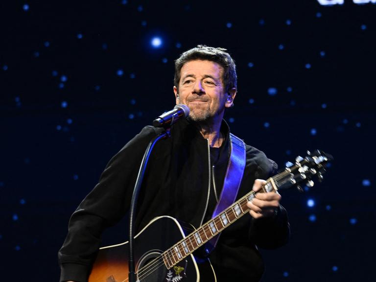 Patrick Bruel steht mit einer Gitarre auf einer Bühne vor dunklem Hintergrund mit sternenartigen Lichtern. 