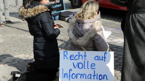 Zwei Frauen knien betend auf dem Boden. Eine hält ein religiöses Buch in der Hand. Eine von ihnen hat ein Schild umgehängt auf dem steht: "Recht auf volle Information".