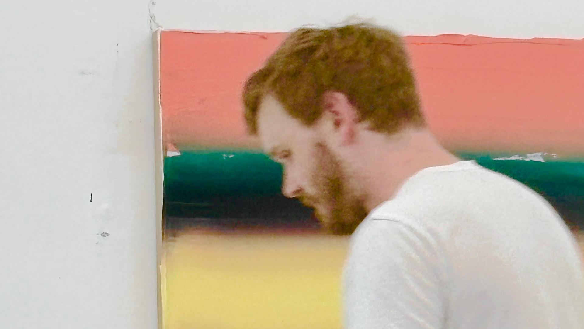 Ein Mann in weißem T-Shirt und mit kurzen rötlichblondem Haar steht vor einem an der Wand hängenden Bild; Felix Becker in seinem Atelier bei der Arbeit 2026