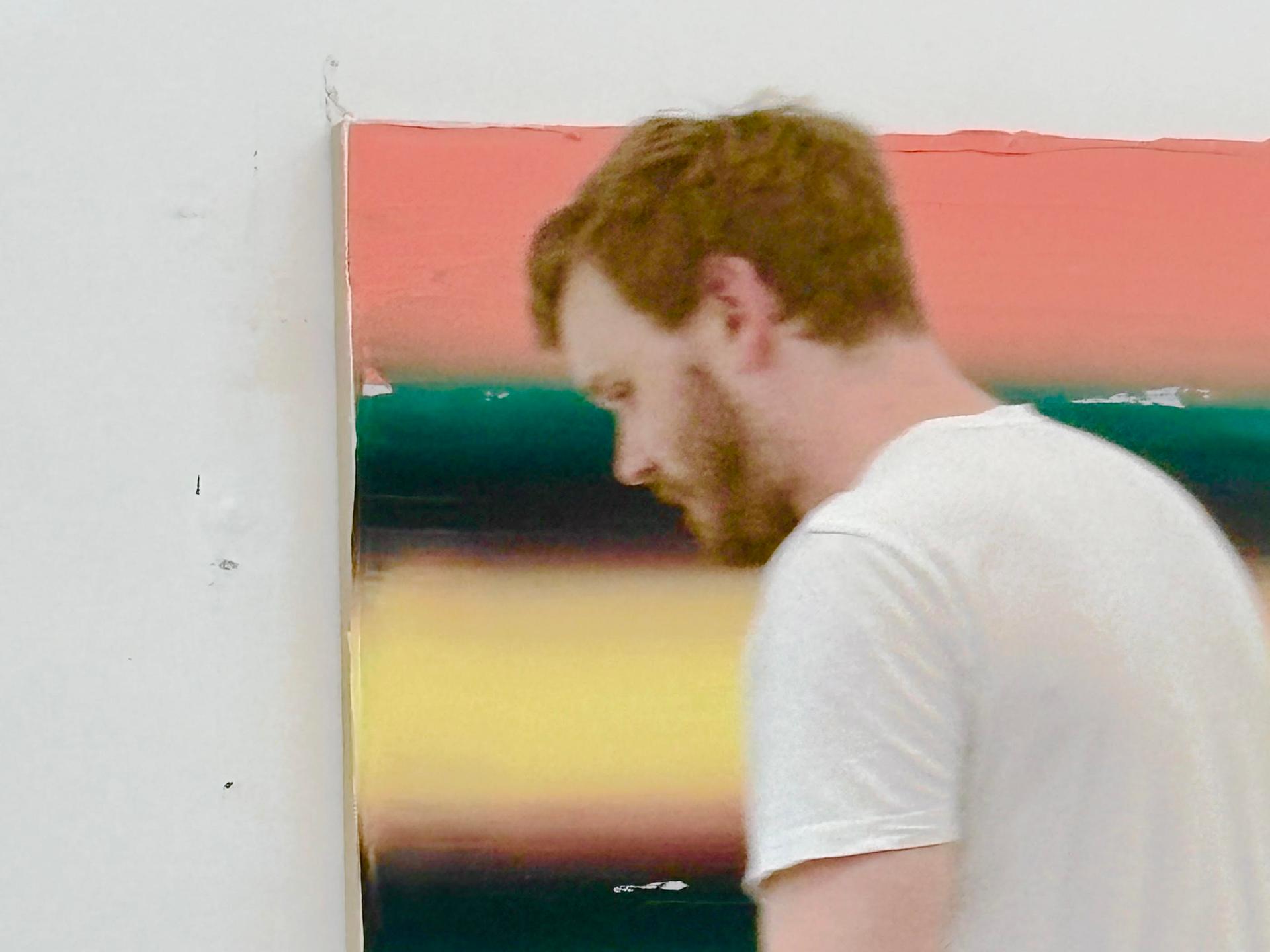Ein Mann in weißem T-Shirt und mit kurzen rötlichblondem Haar steht vor einem an der Wand hängenden Bild; Felix Becker in seinem Atelier bei der Arbeit 2026