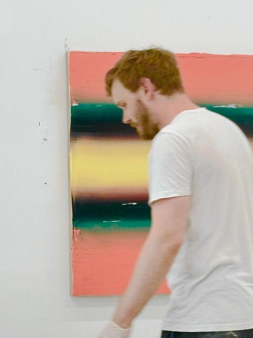 Ein Mann in weißem T-Shirt und mit kurzen rötlichblondem Haar steht vor einem an der Wand hängenden Bild; Felix Becker in seinem Atelier bei der Arbeit 2026